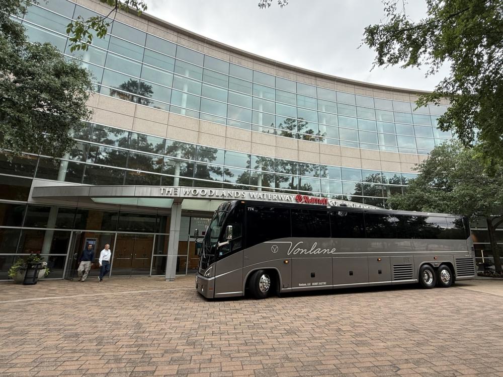 A luxury buses stops in front of The Woodlands Waterway Marriott. The bus is long, silver and black, and has "Vonlane" written in smooth cursive across the side. Behind the bus is a hotel with ground-to-roof windows across the entire frame of the photo.