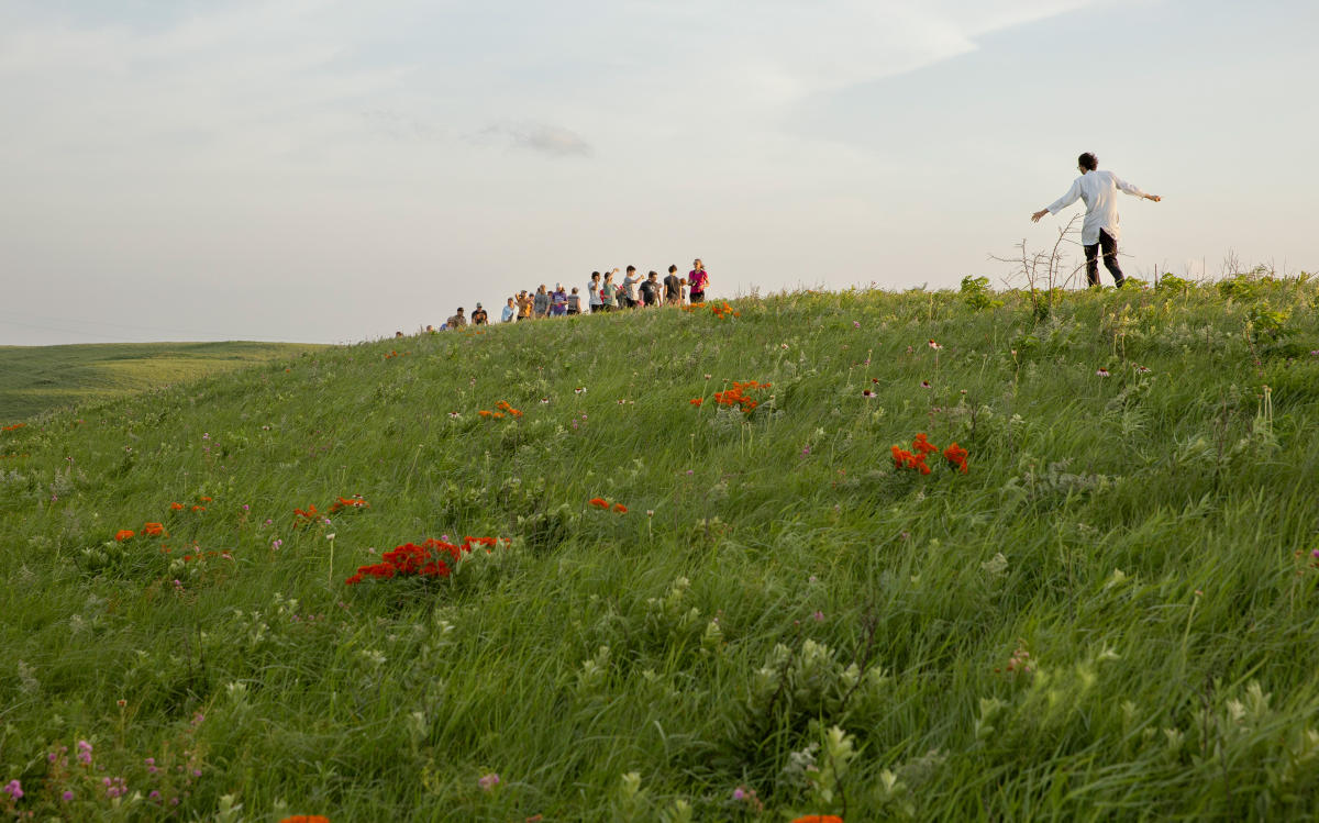a green grass hill with orange wildflowers. People are standing on top of the hill. One man has is arms out to the side
