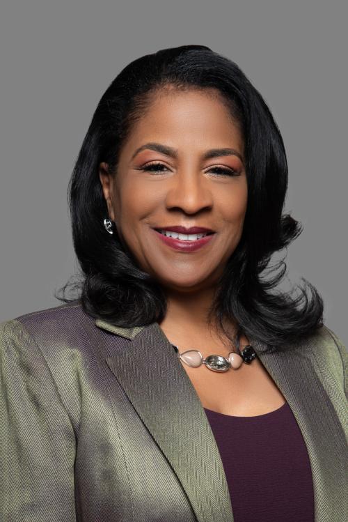 Professional headshot of a woman with black hair, smiling, wearing a grey blazer and brown shirt.
