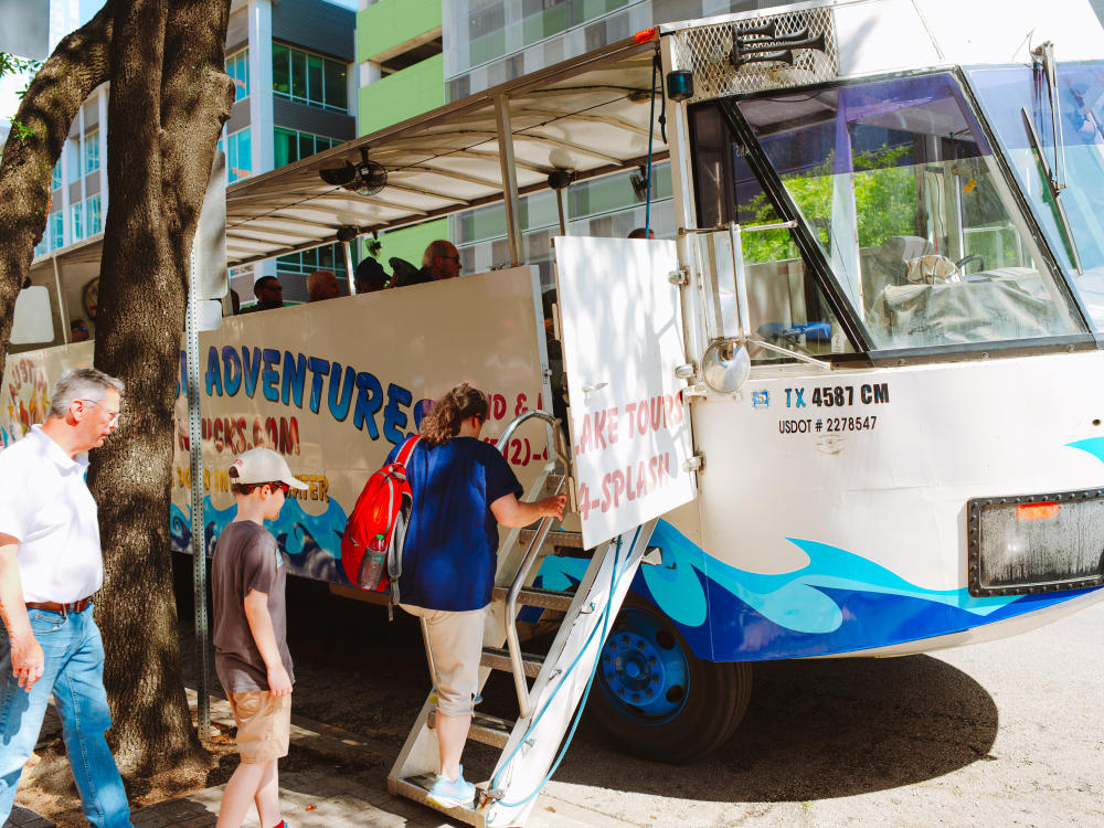 A woman, man and child loading on to the Austin Duck Adventure tour bus, outside of the Austin Visitor Center.