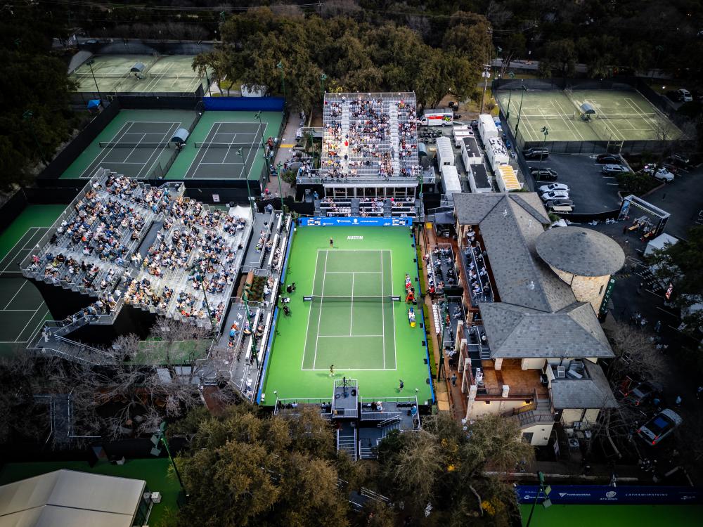 Aerial image of the ATX Open tennis court at Westwood Country Club with a bright green, illuminated court in the middle surrounded by spectator stands with people sitting on bleachers.