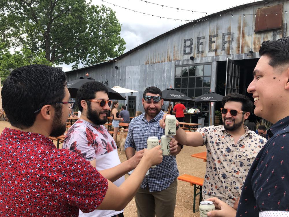 Photo of five young men toasting cans of beer on a Twisted Texas Tour, they are on a patio in front of metal building with sign reading Beer