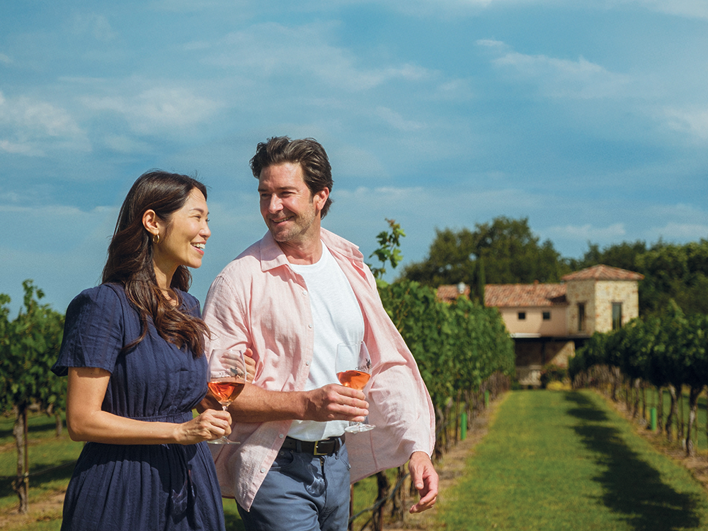 Couple walking through a vineyard with wine glasses in hand on a sunny day.
