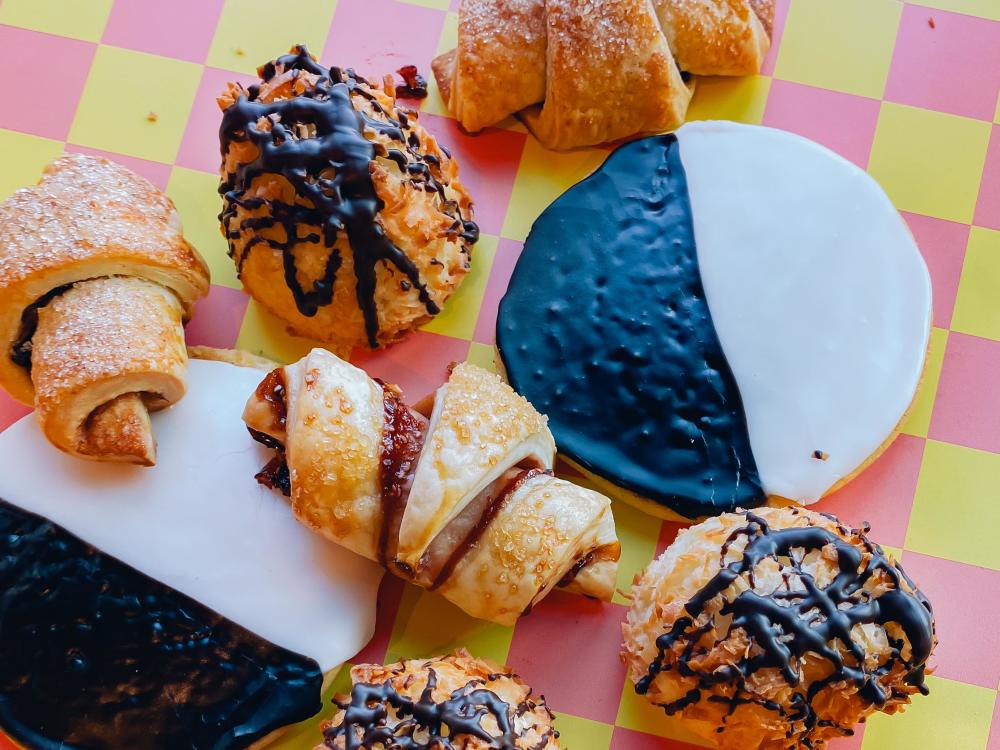 Image of a black and white cookie, coconut macaroon and rugelach sitting on a pink and yellow checkered table.