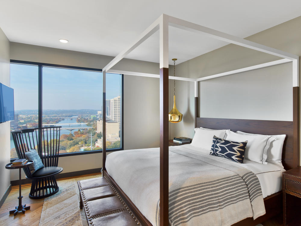 A room at Hotel Van Zandt featuring a large four-poster bed with white and grey linens and a chair in the corner overlooking a window with a view of Lady Bird Lake.