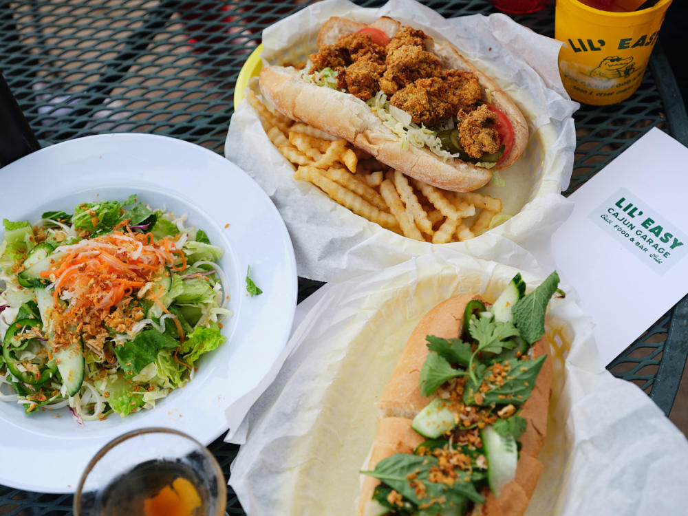 Three different dishes, two different po'boys and a green salad, sitting on a patio table surrounded by a menu and cup reading "Lil' Easy".