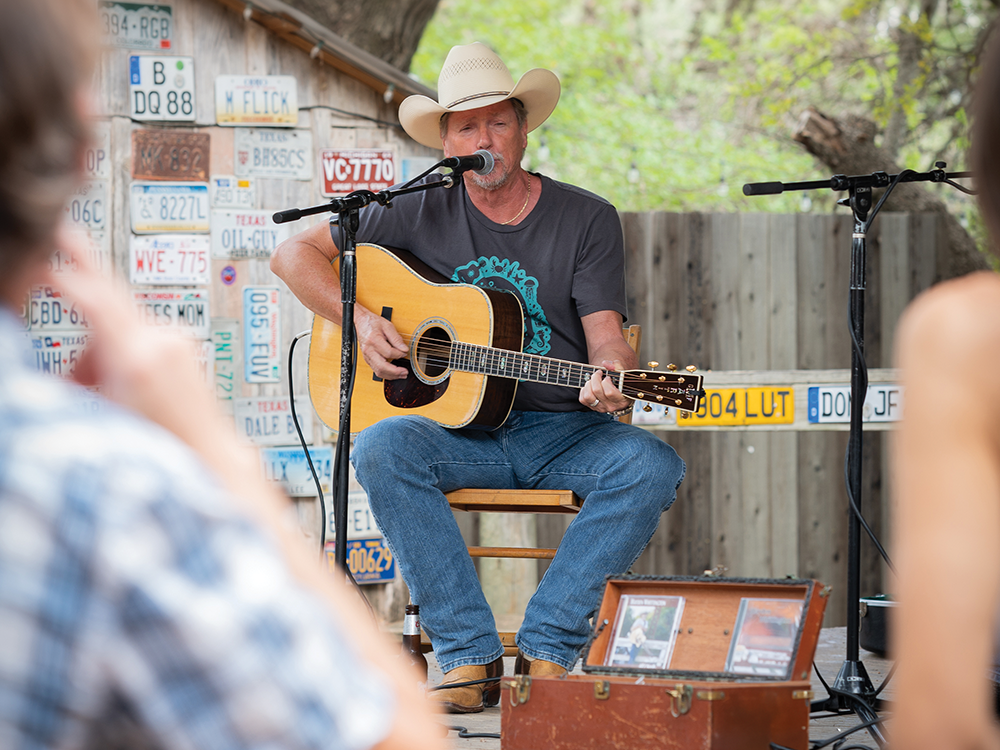 Man in a cowboy hat and cowboy boots playing guitar and singing into a microphone on a stage covered in old license plates.