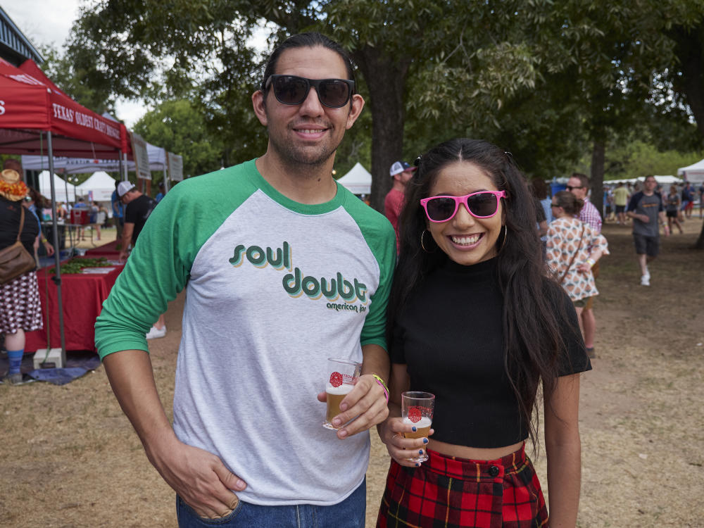 Man wearing green and white baseball tshirt reading Soul Doubt American IPA holding a beer. To his left is a woman in a black shirt and red and black plaid skirt holding a beer. They are both wearing sunglasses and smiling at the camera