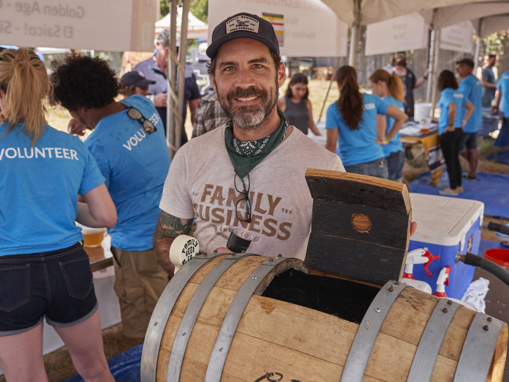 Man wearing a shirt reading Family Business Beer Company standing behind an oak barrel keg. Festival volunteers in blue shirts stand behind him