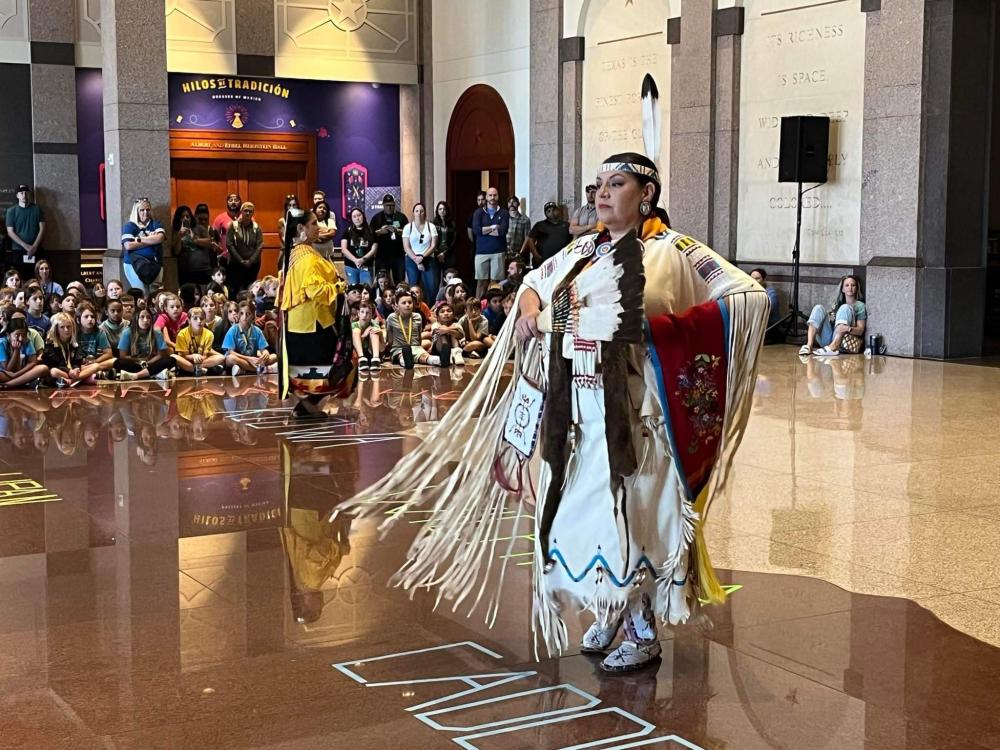 Native American woman in traditional dress, in front of crowd of students.