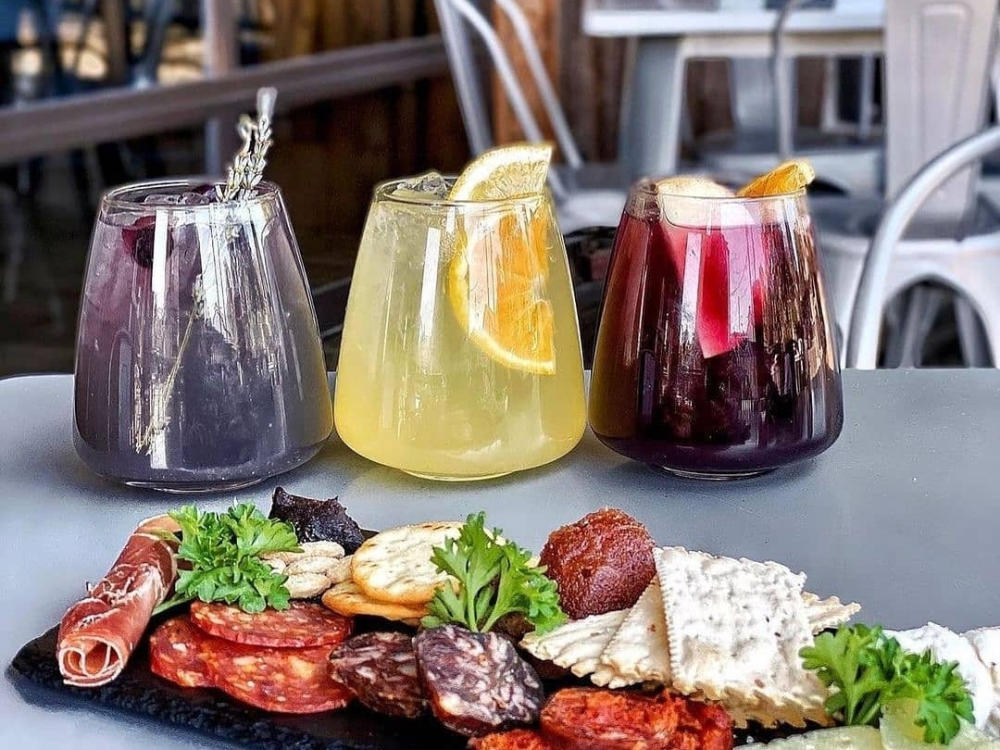 photo of three colorful cocktails sitting on a table behind a plate of assorted cheeses, crackers and cured meats at Botero Tapas + Wine Bar in Boerne Texas