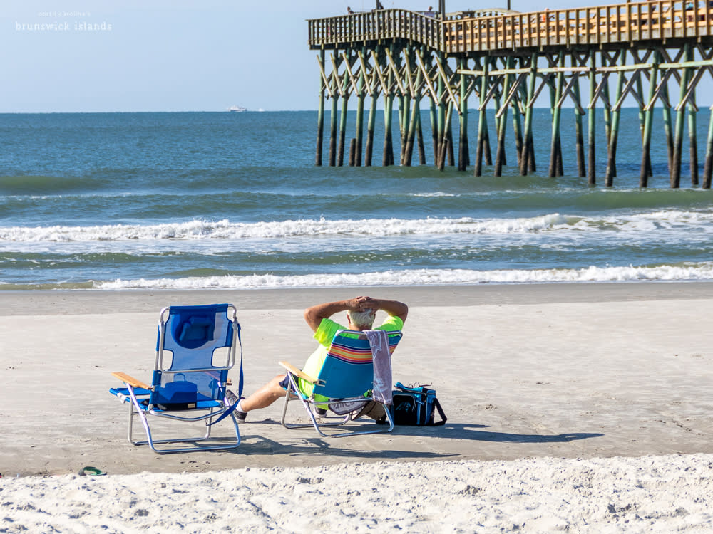 man leaning back in a beach chair on the beach with an ocean pier extending in the background
