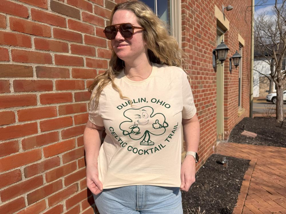 A woman with long blonde hair and sunglasses poses against a brick wall, wearing a beige T-shirt that reads 'Dublin, Ohio Celtic Cocktail Trail' with a smiling shamrock holding a cocktail.