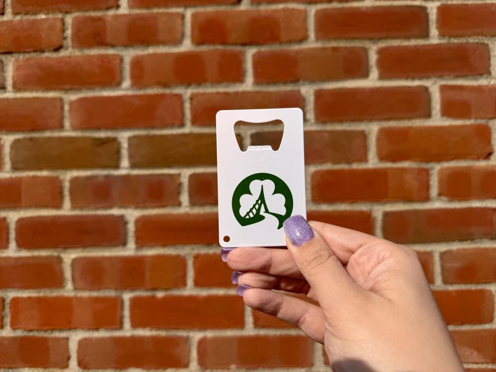 A hand with purple nail polish holds a white bottle opener featuring a green shamrock logo with a bridge design, against a red brick wall background.