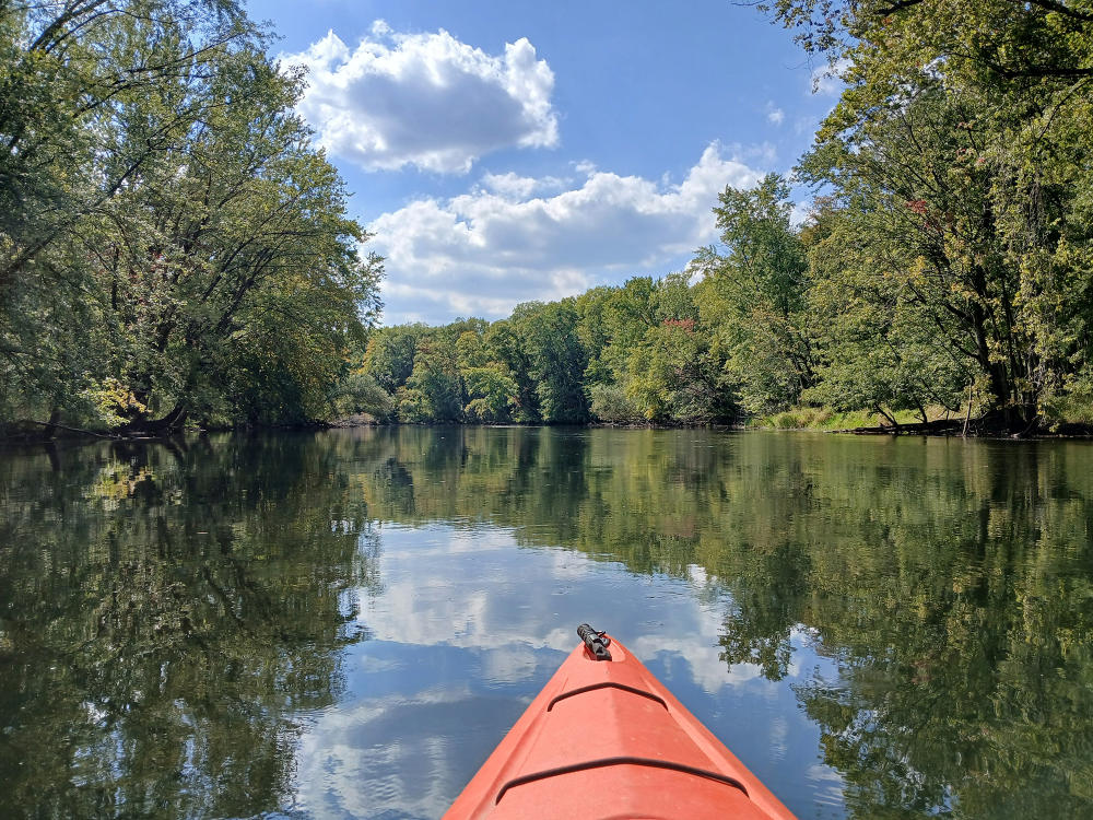 An orange kayak sits in the middle of the Kalamazoo River, with trees surrounding the banks.