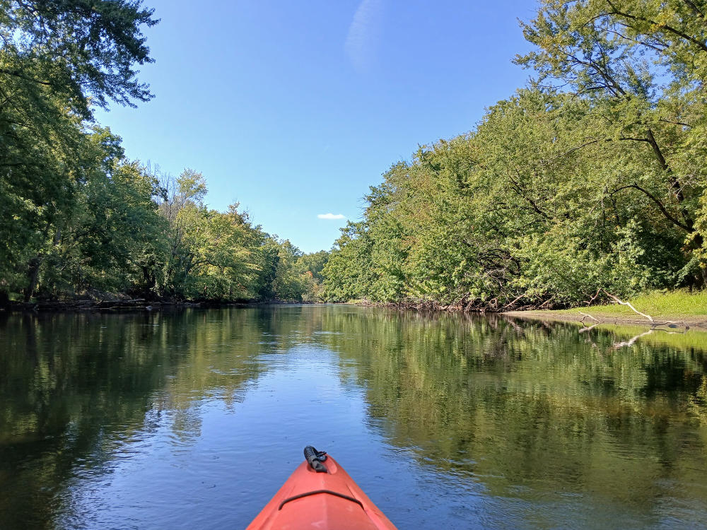 An orange kayak sits in the center of the Kalamazoo River, surrounded by banks of Green trees, as if kayaks from Battle Creek to Augusta.