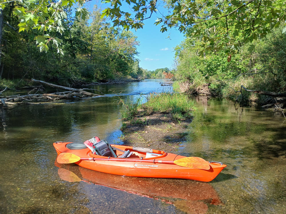 An orange kayak sits partially in the Kalamazoo River and partially on the shore line with its paddle displayed on top of the kayak.