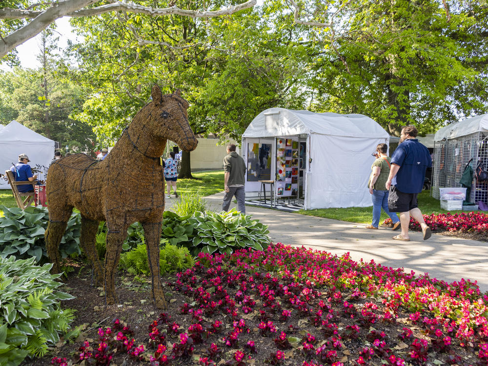Red roses surround a grass sculpture of a horse while two people walk on a sidewalk behind the structure. There are tents along the sidewalk selling local artist works.