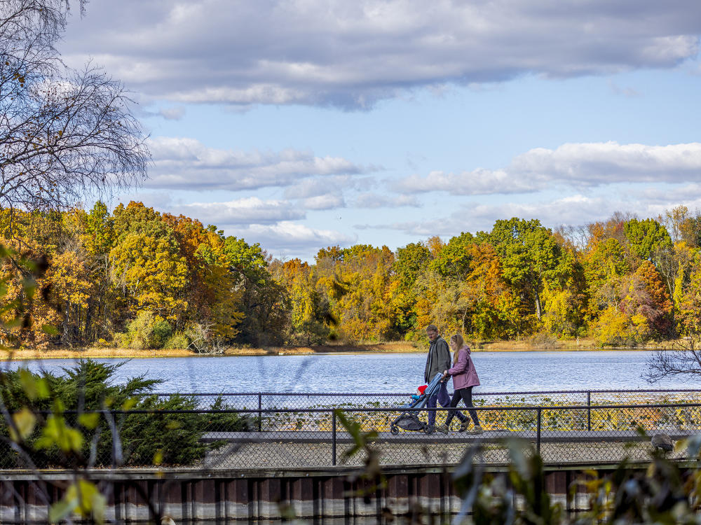 A couple with a toddler in a stroller walk on a path at the W.K. Kellogg Bird Sanctuary that overlooks the water with fall-turning trees in the background.