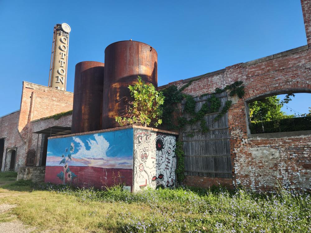 old brick wall with murals and rusted water towers on top and a vertical sign in the background that says "Cotton Mill" on it