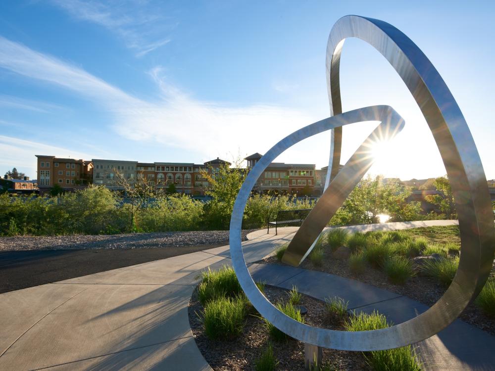 Large metallic sculpture with intertwined loops stands in a park, illuminated by the setting sun