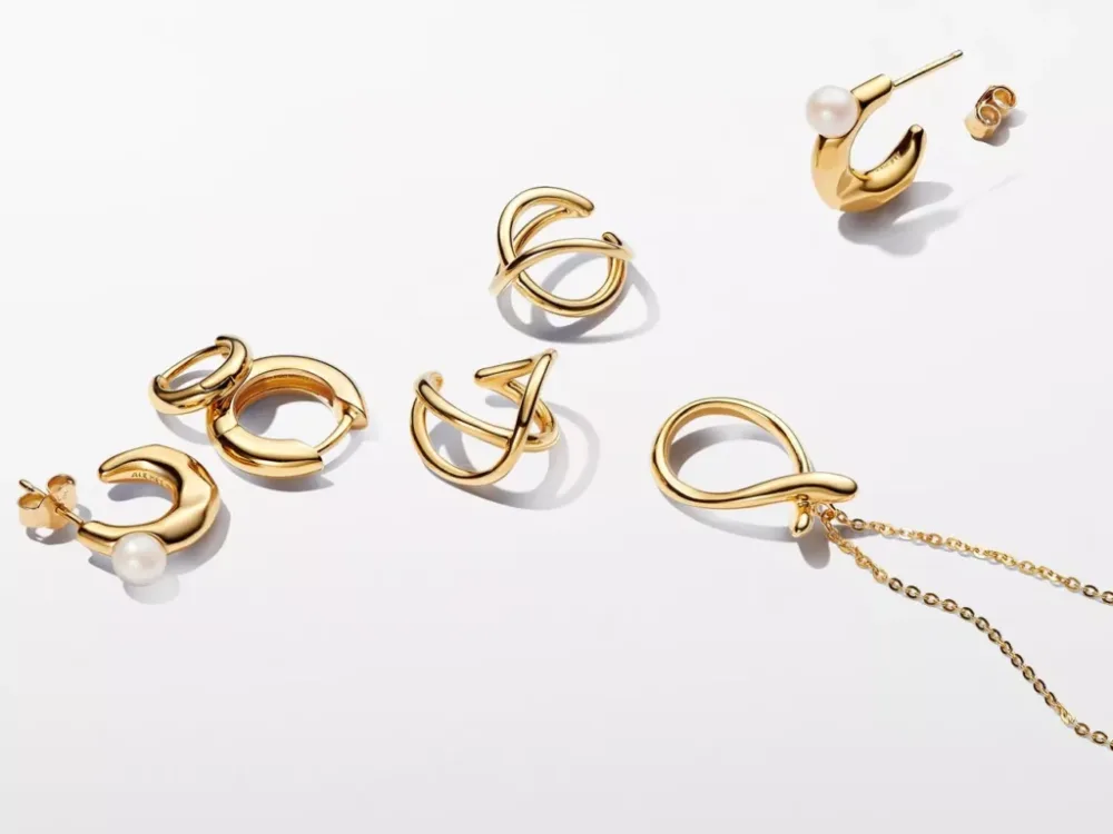A collection of modern gold jewelry pieces, including hoop earrings, a pearl earring, and a gold necklace with a looped pendant, displayed on a white background.