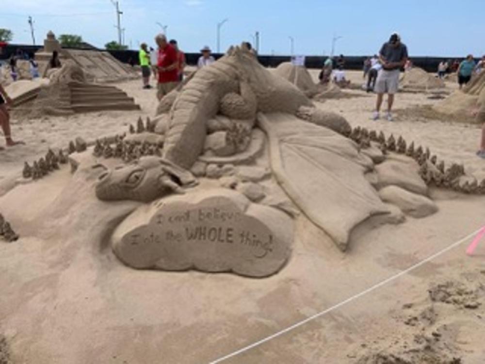 Sand sculpture of a dragon lying on some rocks. In front is a cloud-shaped structure that says, "I can't believe I ate the whole thing."