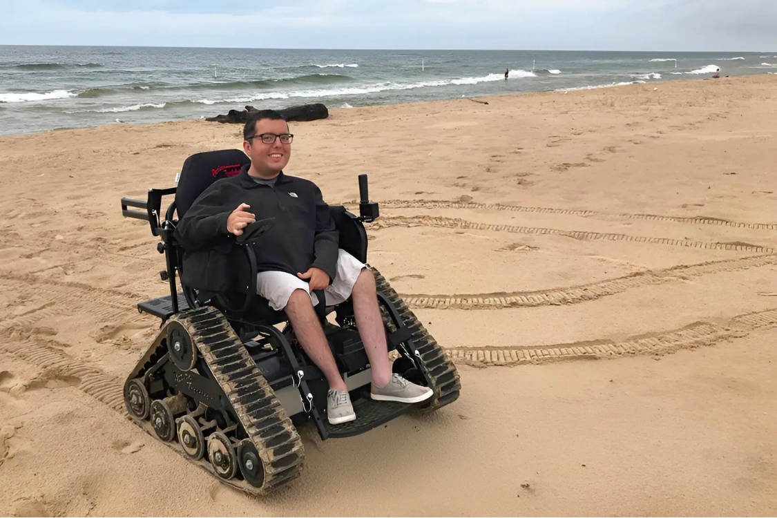 Person in a motorized all-terrain wheelchair smiles on a sandy beach, with Lake Michigan in the background.