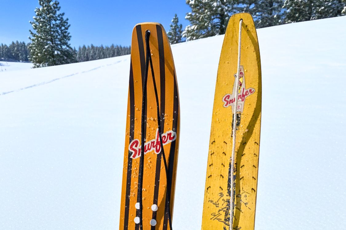 Pair of Snurfer Boards, one orange and one yellow, standing upright in snowy landscape under a clear blue sky.