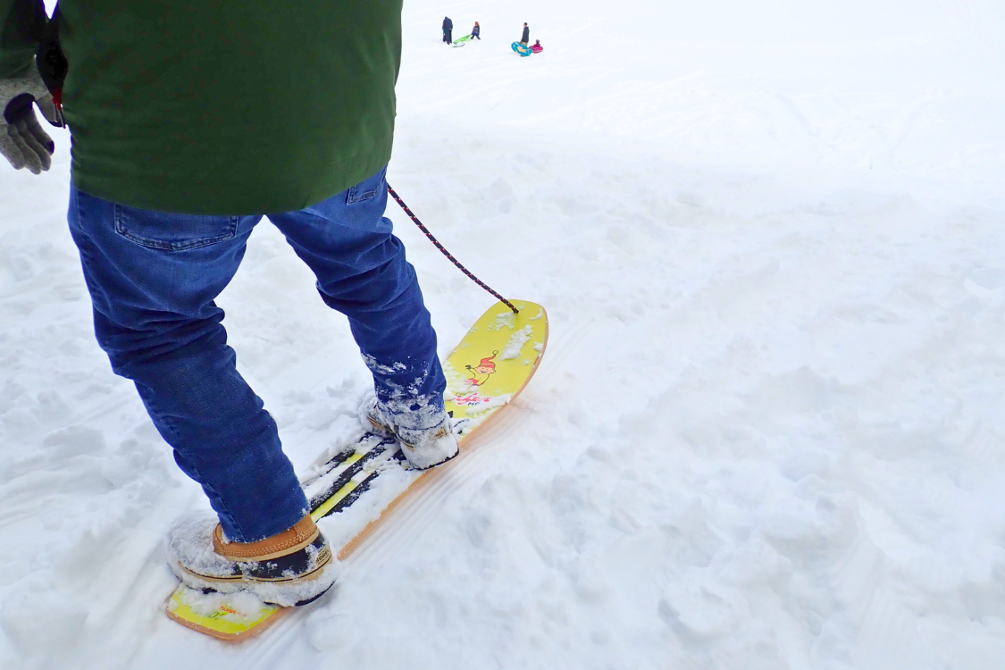 A person stands on a yellow snowboard in the snow, with other people sledding in the background.