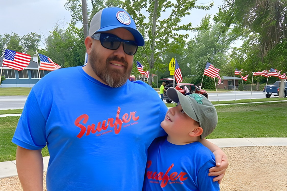Two people wearing blue t-shirts and caps stand together in a park with multiple American flags in the background.