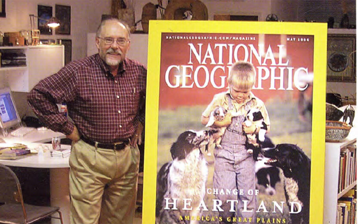 a man standing in his office next to a magazine cover printed very large. The cover has a boy surrounded by dogs on it