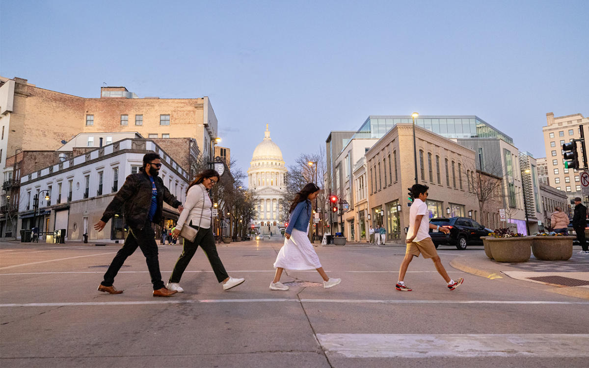 A family of four visiting Madison, WI walks across the street with the Wisconsin State Capitol and art museum in the background