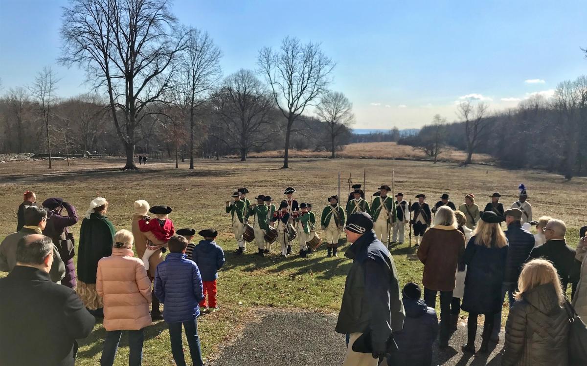 A crowd looks on during a reenactment featuring soldiers lined up and dressed in green and white clothing.