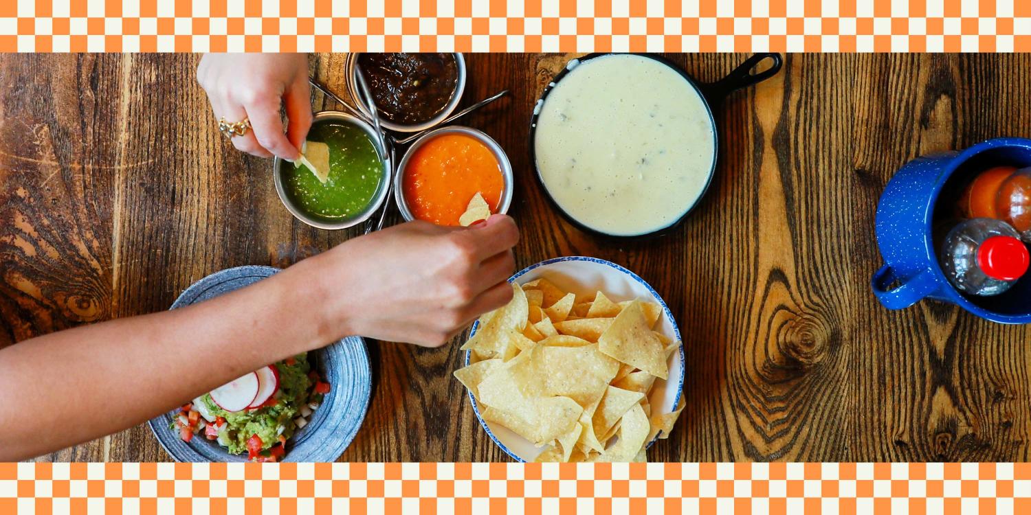 A person dips a tortilla chip into a bowl of salsa near guacamole, queso dip, and various sauces on a wooden table.