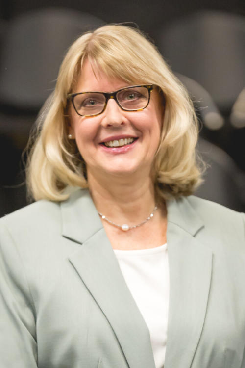 Professional headshot of a woman, with blonde hair, black glasses, smiling, with a sage green blazer.