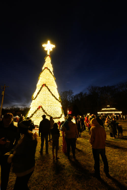 Visitors gather around a towering Christmas tree illuminated with thousands of white lights and topped with a glowing star during Light Up Charlestown in Charlestown, Indiana, with festive holiday displays lighting up the night.