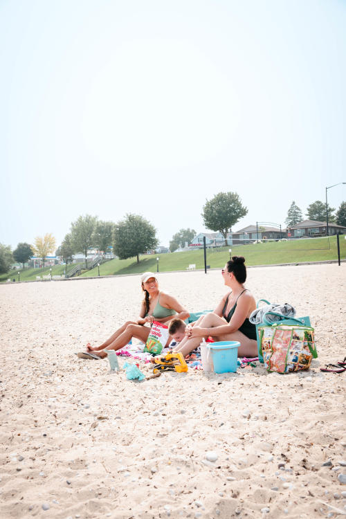 Two adult women and a young boy sitting on the beach at Crescent Beach in Algoma, Wisconsin. They are snacking on chips and playing with sand toys.