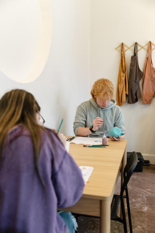 Two people painting ceramic pieces during a hands-on pottery session at Bisque by The Vessel at Stovehouse. Paintbrushes, glaze bottles, and art supplies sit on the table in a bright studio space with aprons hanging on the wall.