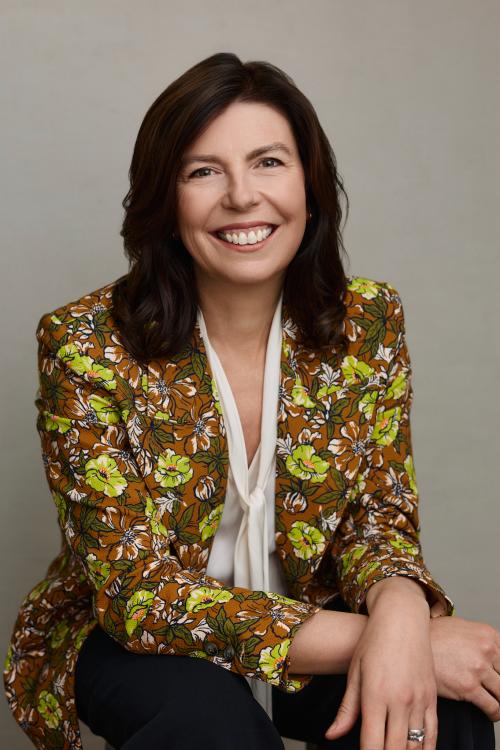 Headshot of Leslie Bruce. She's sitting, smiling at the camera with her hands crossed. She's wearing a white blouse and a floral-print blazer.