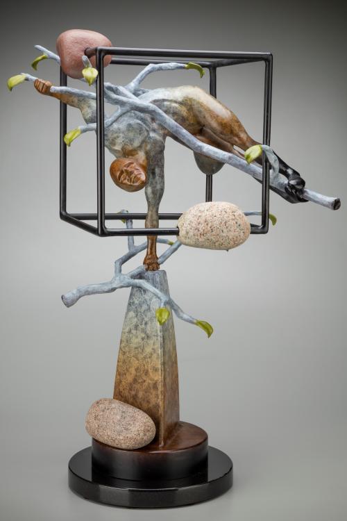 The camera faces a sculpture by Thomas Wargin. It is an abstract piece featuring a contorted male figure encased in a cube made of thin, black, metal bars. The figure, balanced on a stone pillar with his right fist, clings to a tree limb with his left hand and feet. Other natural elements like leaves and smooth river stones are also incorporated.