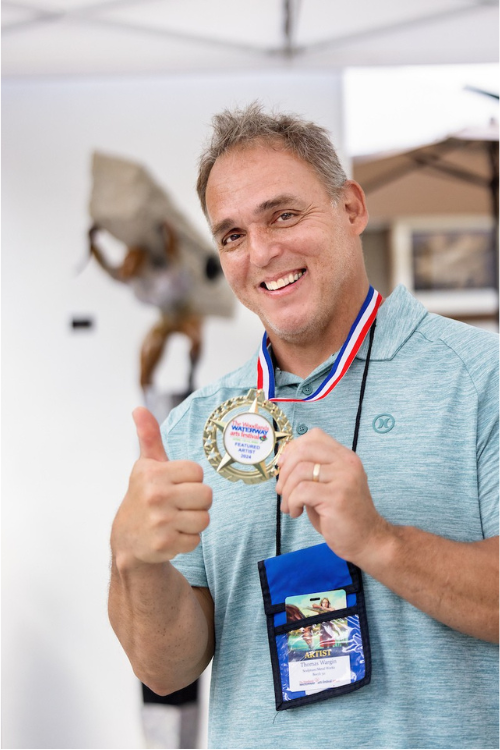 A man, 40s-50s, smiles and offers a thumbs up to the camera. He is wearing a light blue shirt, a dark blue lanyard carrying his name tag, and a star-shaped medal on a red, white, and blue-striped lanyard. Blurred in the background is one of the man's sculptures, a figure struggling to carry a rough, concrete pillar on his back.