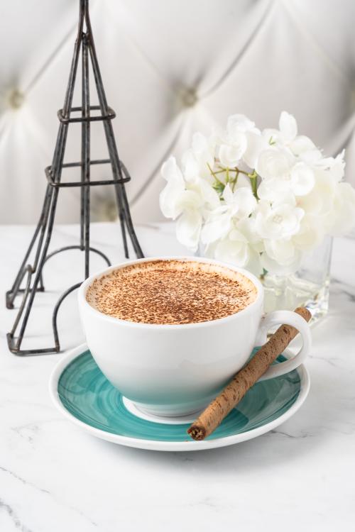 A close-up shot of a tiramisu latte, lightly dusted with cinnamon. A cinnamon stick is propped up through the teacup handle. Behind the cup are (fake) white flowers and a black silhouette of the Eiffel Tower.