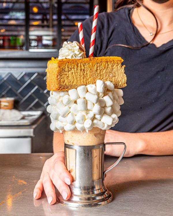 Image of a milkshake served in a glass. Milkshake is topped with a marshmallow rim and a whole slice of pumpkin cheesecake.