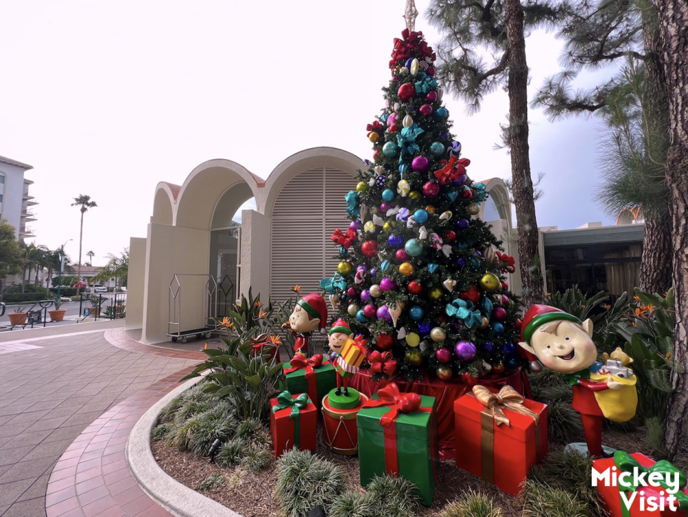 Image of a large Christmas tree decorated with gold, blue, and red ornaments. Christmas presents wrapped in red and green wrapping paper can be seen under the tree. The tree is placed outside, right next to the entrance of the Howard Johnson Anaheim Hotel & Water Playground.