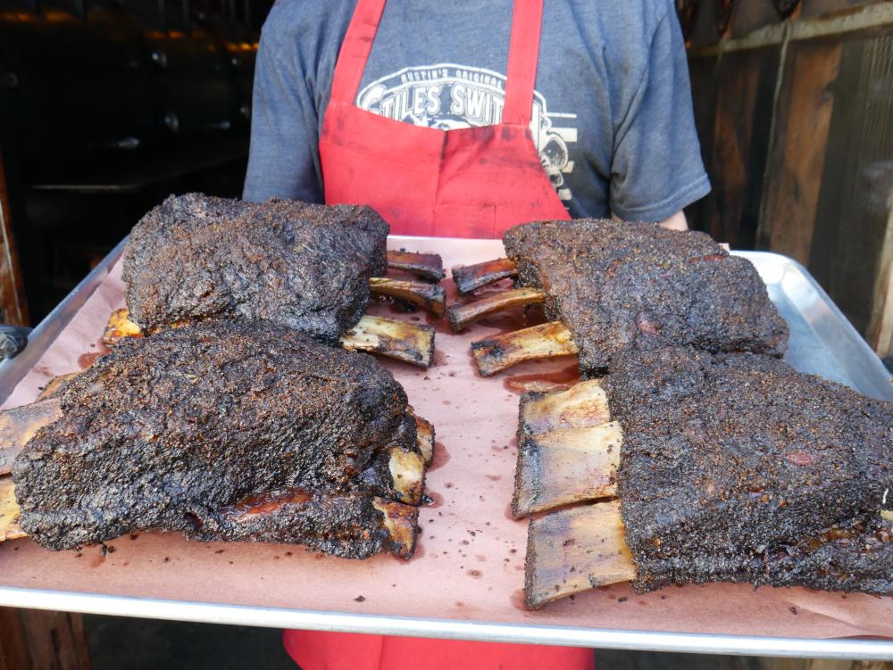 Photo of a pitmaster holding a tray with huge smoked beef ribs.