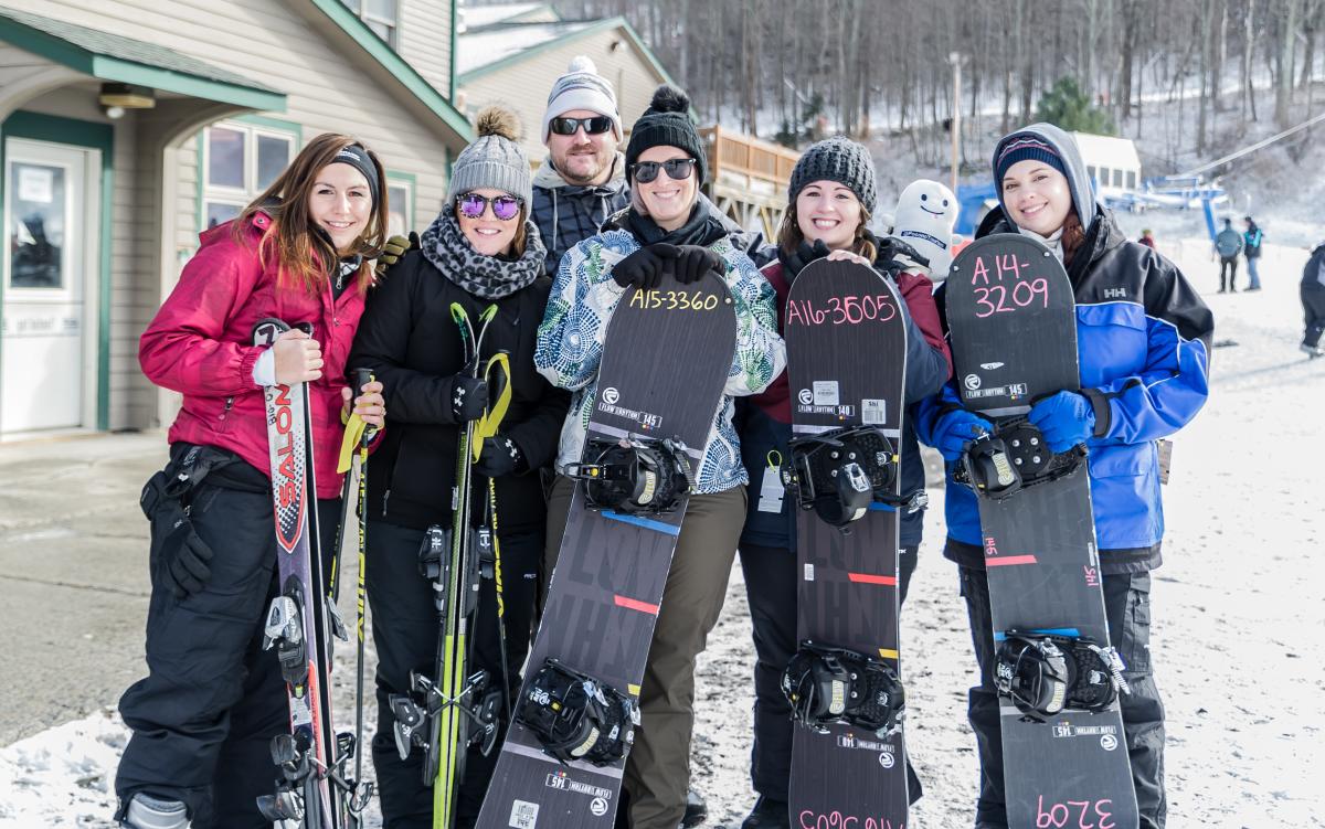A group of five women and one man pose with their snowboards outside a ski lodge