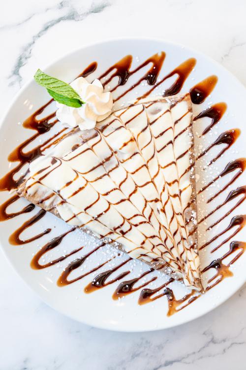 A close-up shot of a tiramisu crepe coated in cream cheese, drizzled in chocolate syrup, and garnished with whipped cream and a mint leaf.