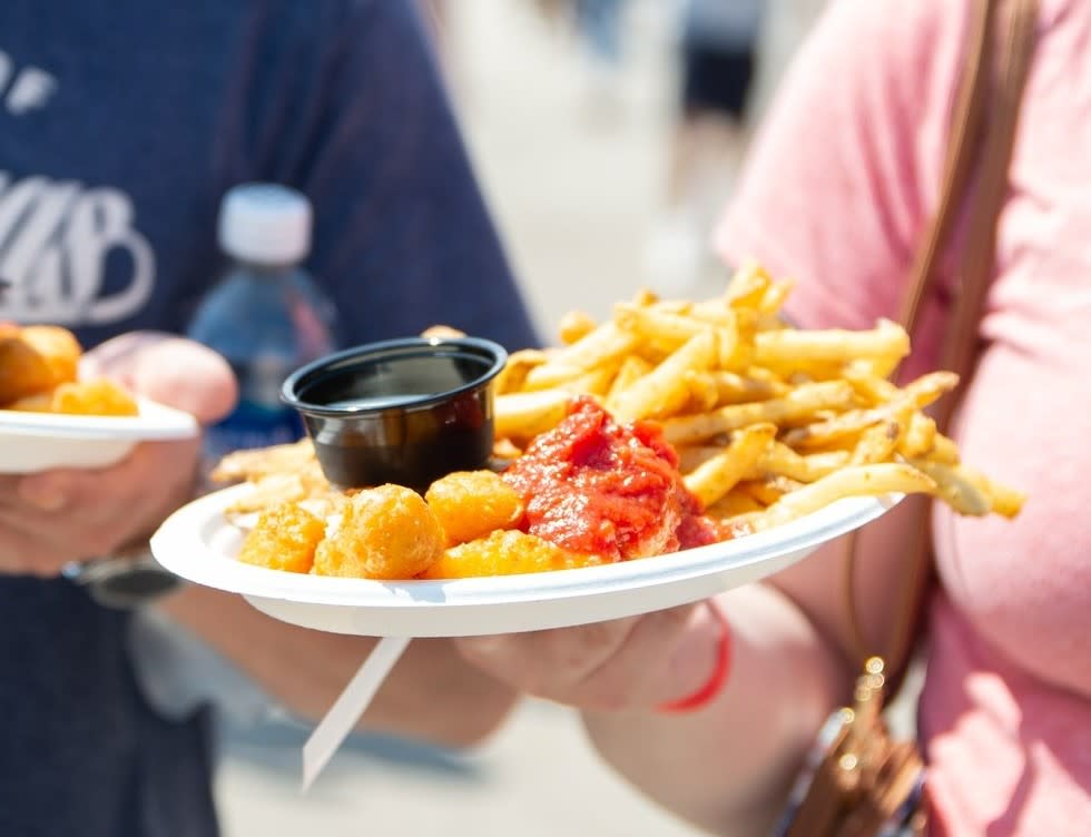 A close-up of a person holding a paper plate filled with golden French fries, cheese curds, marinara sauce, and a small black cup of dipping sauce at an outdoor festival. Another person holding a similar plate and a water bottle is visible in the background, slightly out of focus.