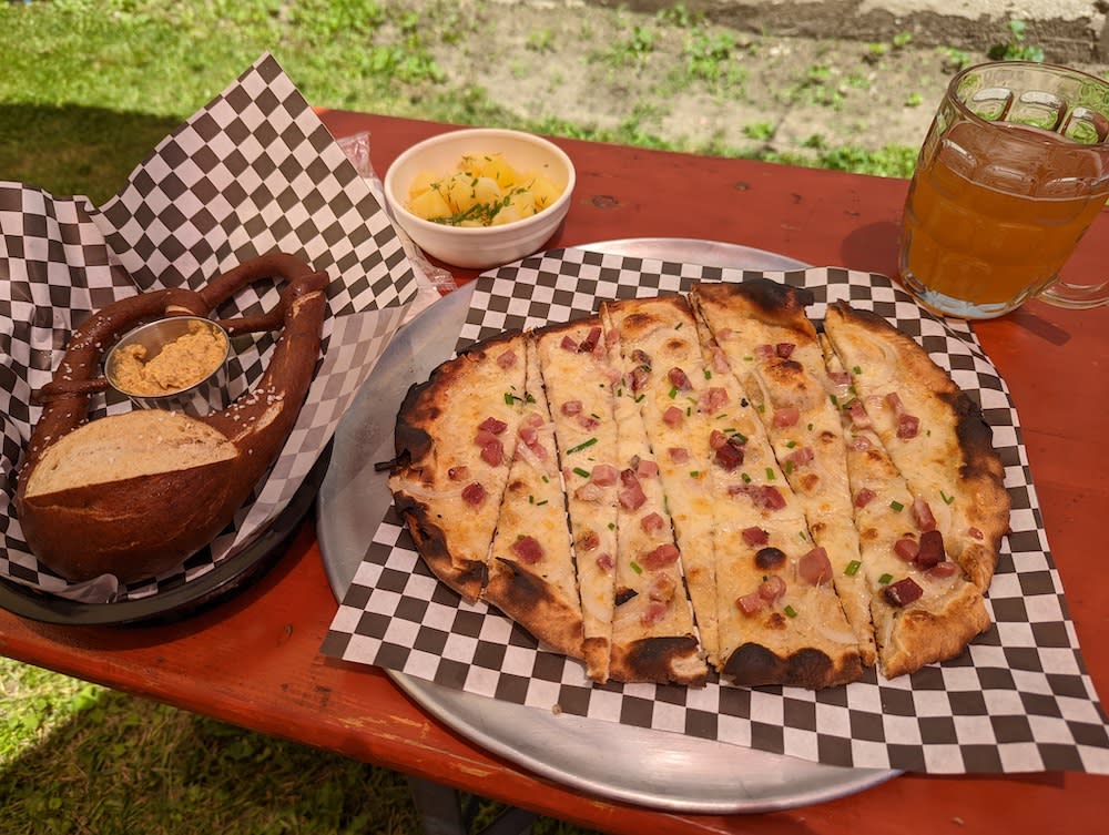 A soft pretzel and German flammekuchen sit on a red picnic table with a pint of beer at Tuba Baking Co
